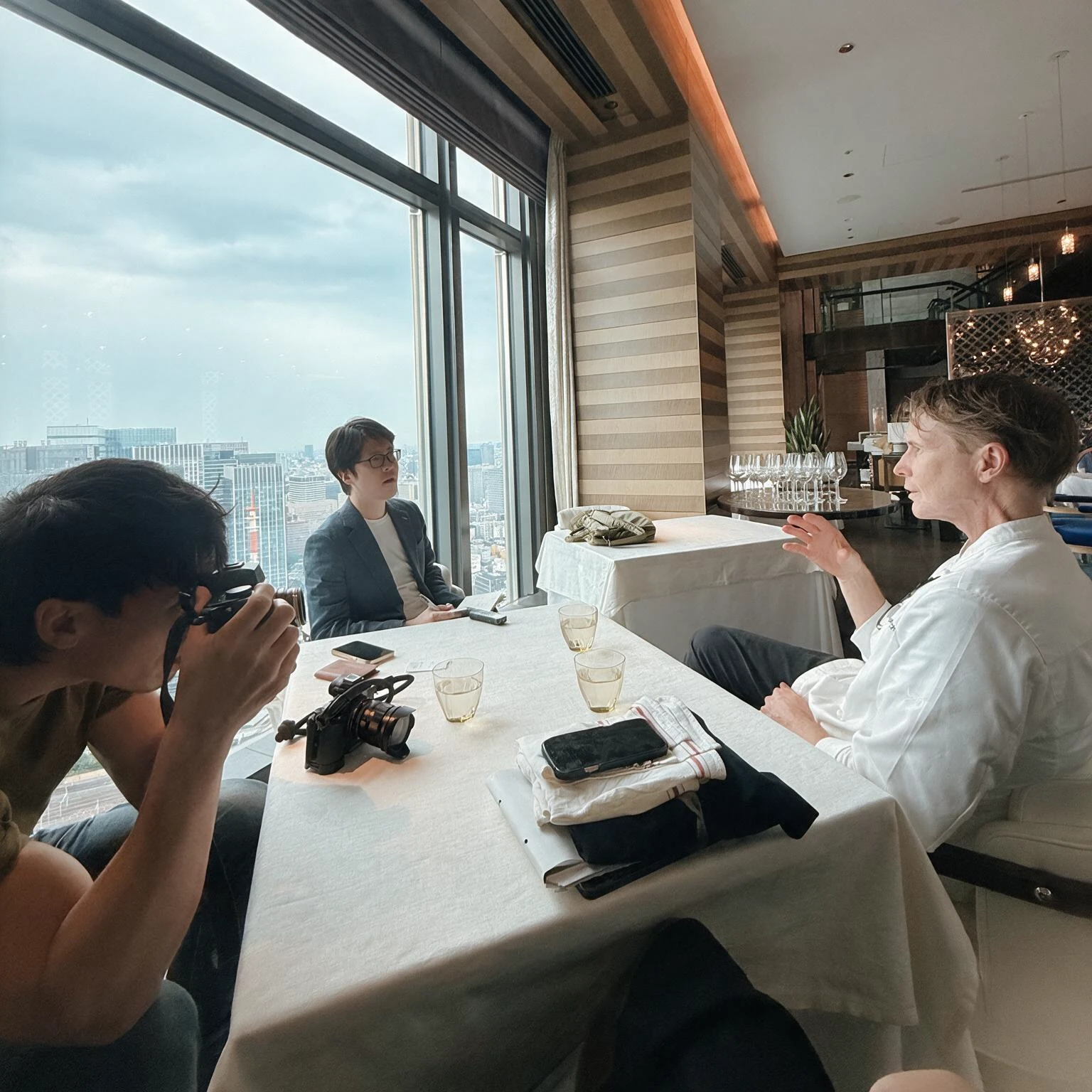 A photographer and an interviewer at a restaurant table with a chef in kitchen whites